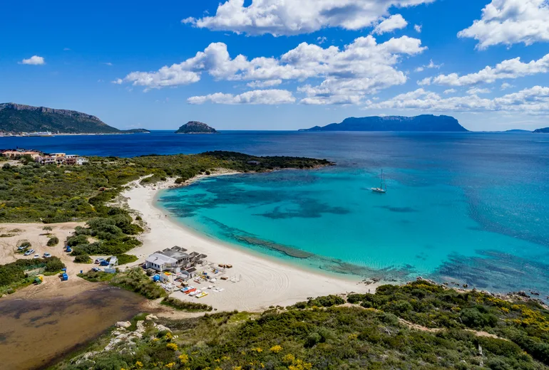 Aerial of a white sand crescent beach with a single sailboat anchored over pale shallows near Golfo Aranci, Sardinia