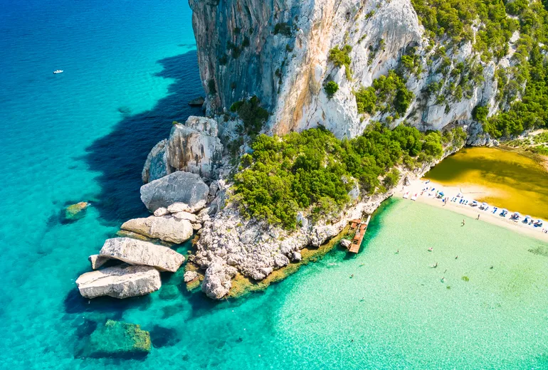 Aerial of limestone cliffs, motorboats and a turquoise beach cove showing pale sand and darker seabed patches, Sardinia