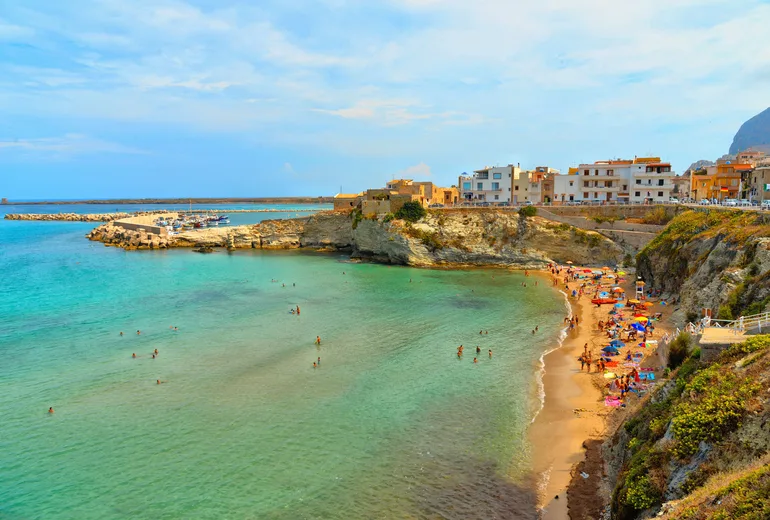 Bathers on a golden beach beside a small Sicilian town with a stone fort