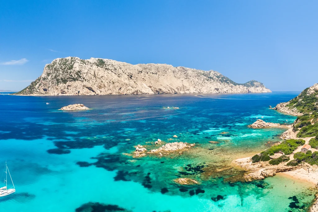 Aerial panorama of Tavolara island with sailboat anchored in turquoise shallows, Sardinia