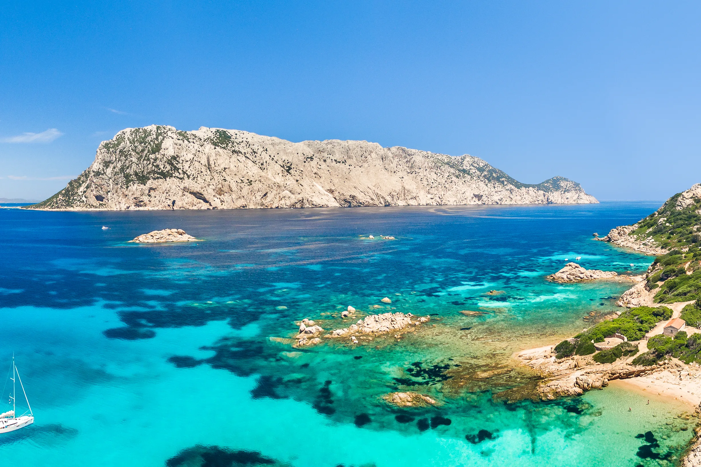 Aerial panorama of Tavolara island with sailboat anchored in turquoise shallows, Sardinia