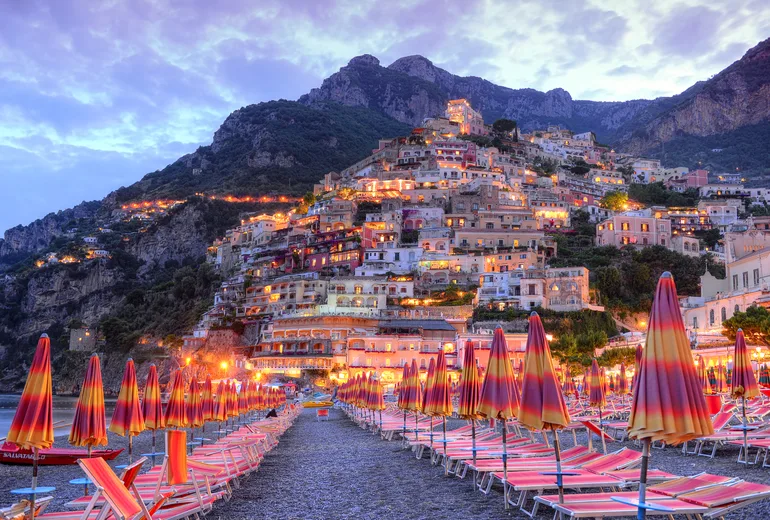 Positano hillside village glowing at dusk above rows of closed beach umbrellas