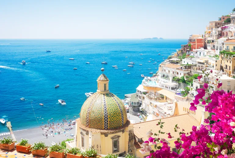 Positano majolica church dome, bougainvillea and busy beach with boats offshore, Amalfi Coast