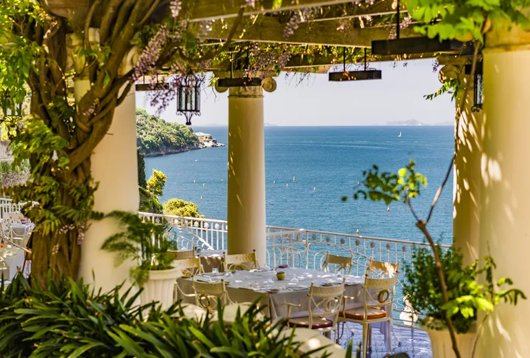 Restaurant terrace under a wisteria-covered pergola with columns overlooking the sea