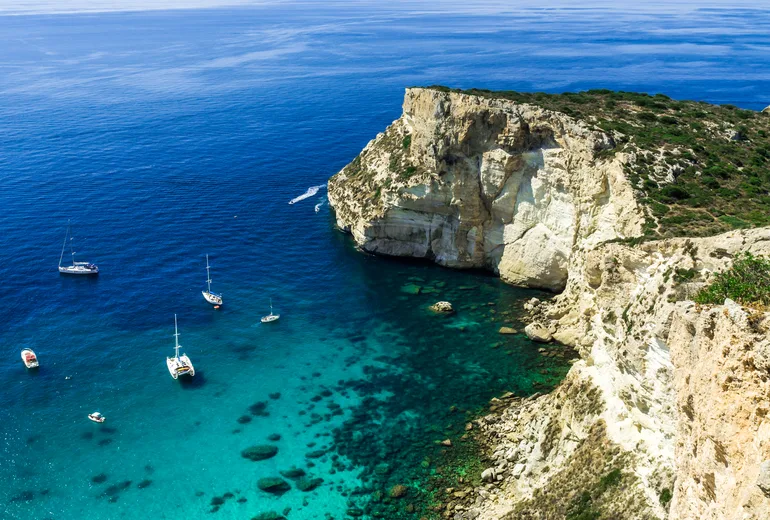 Sailboats and a catamaran anchored beneath a white limestone headland in a turquoise cove