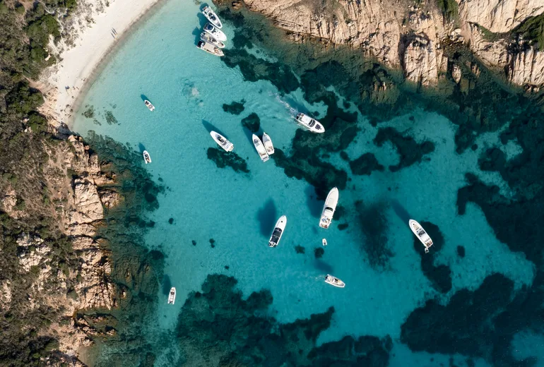 Top-down aerial of boats anchored in a turquoise rocky cove with a white sand beach, Sardinia