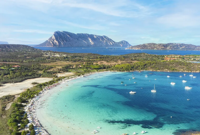 Vertical aerial of a busy turquoise bay with anchored boats and Tavolara island behind, Sardinia