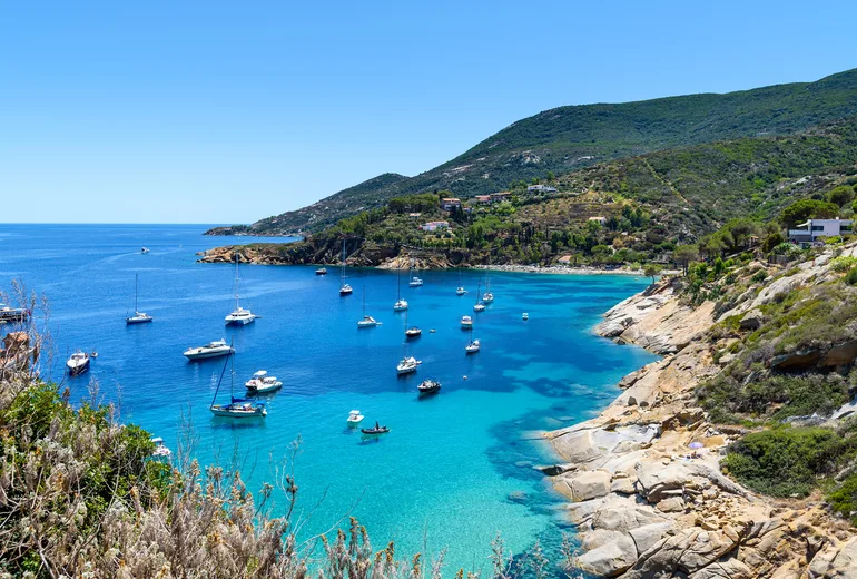 Yachts and sailboats anchored in a turquoise cove off a hilly coastline
